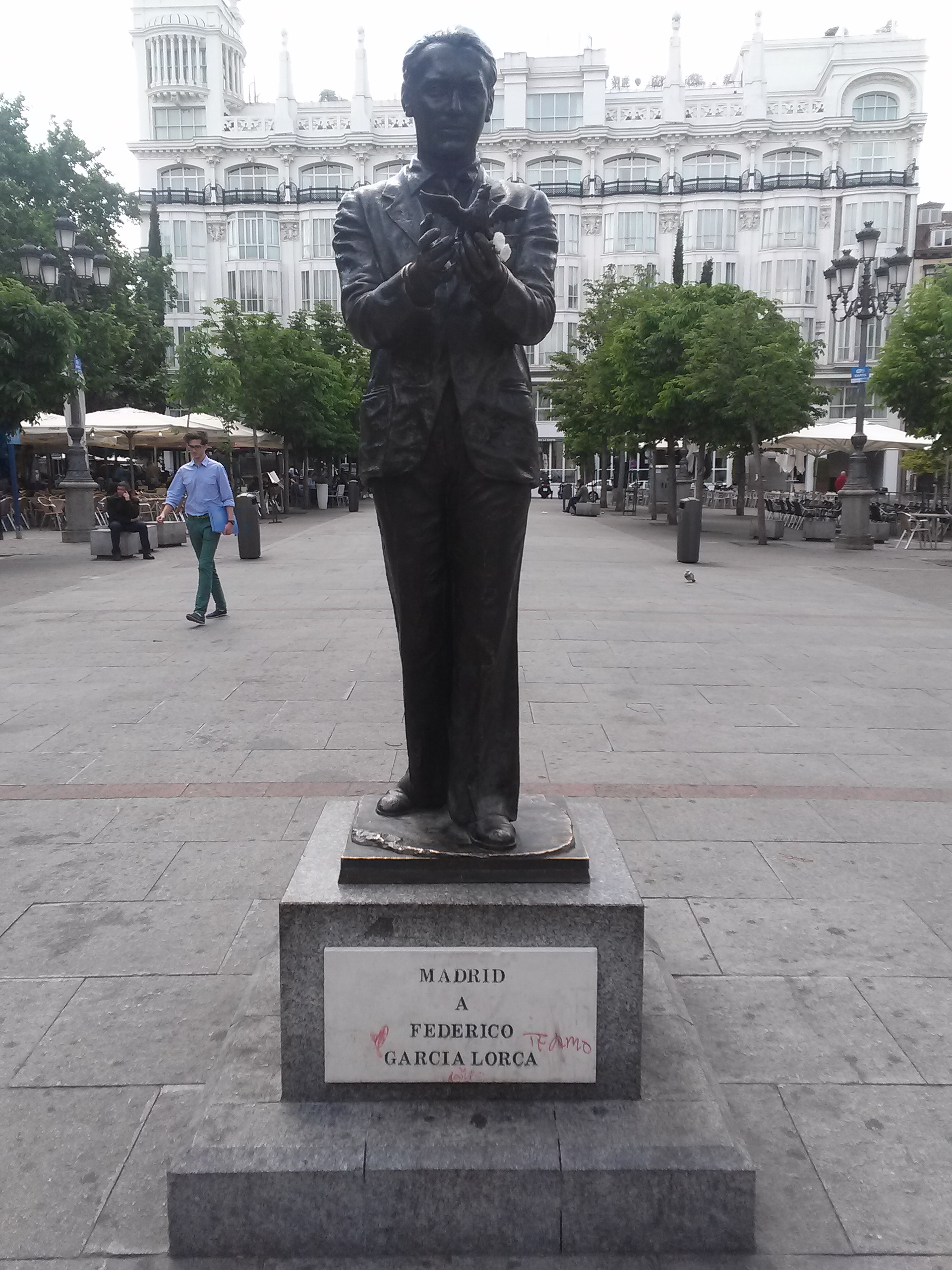 A photo of the statue of poet Federico Garcia Lorca in Plaza de Santa Ana, Madrid. The statue depicts Garcia Lorca holding a lark; visitors have placed flowers in his hands.